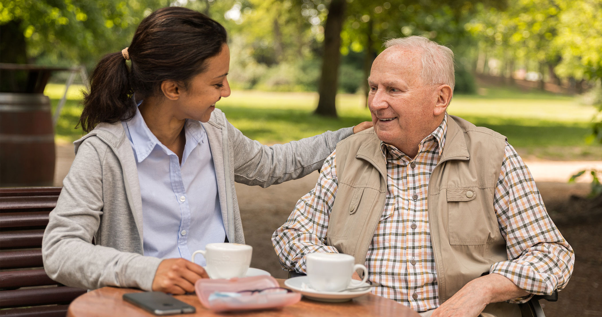 happy caregiver sitting outside with senior man sitting and drinking coffee